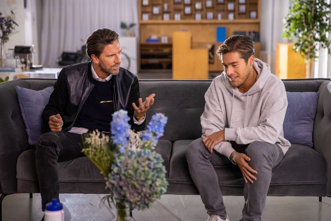 Two men sitting on a couch talking to each other.